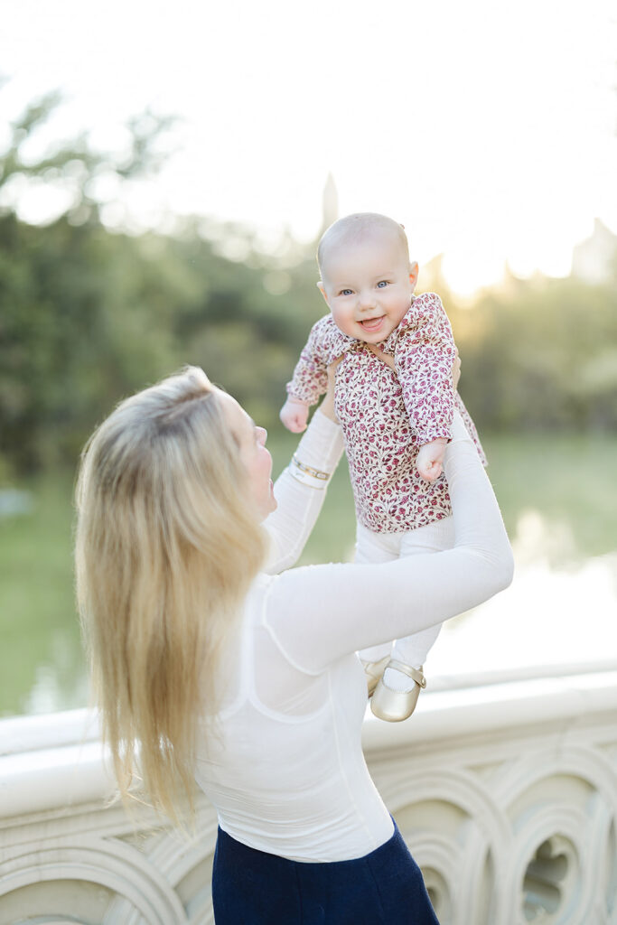 Mom holding her laughing baby girl on Bow Bridge, documented by an NYC family photographer in Central Park, Heirloom by Melissa Photography