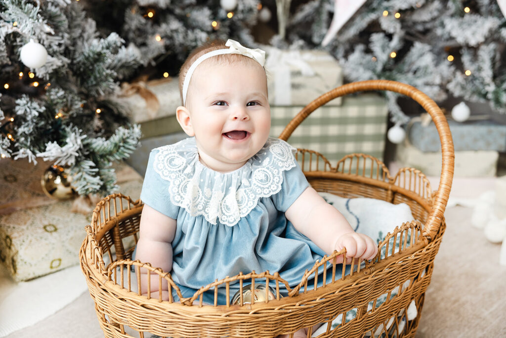 Baby's first holiday photos NYC, baby girl in blue velvet and lace laughing in a wicker bassinet at Heirloom by Melissa, Flatiron District