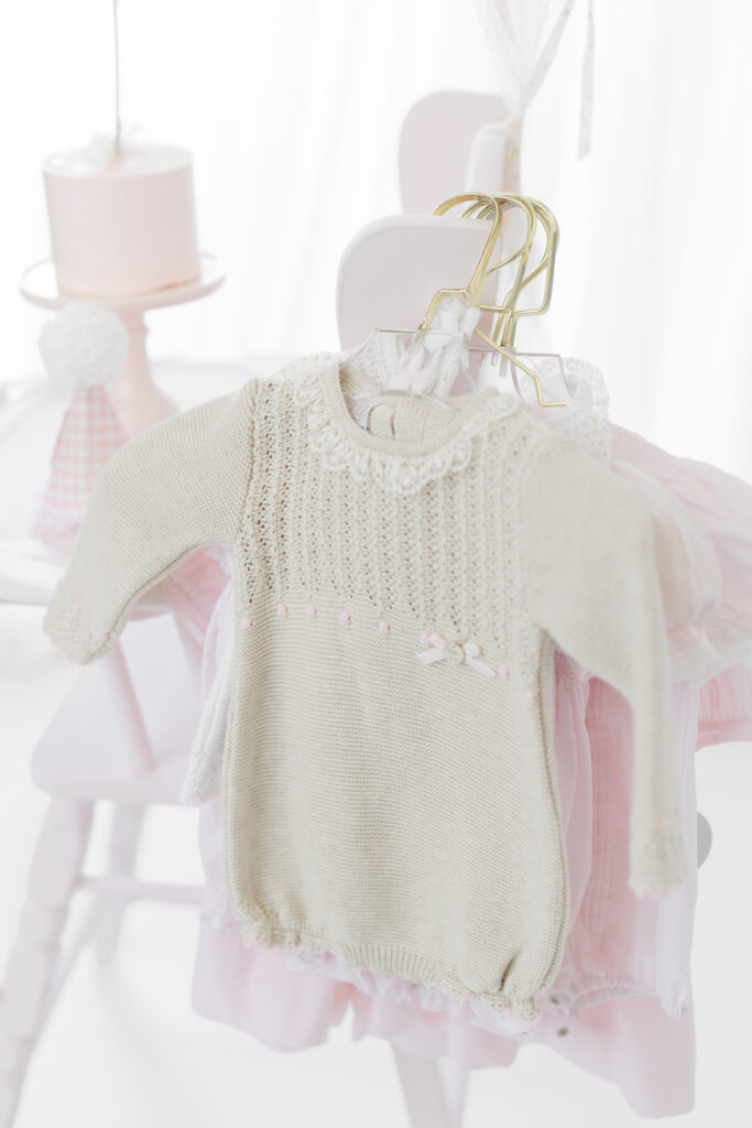Pink and cream first birthday outfits hanging beside Eleanor’s cake