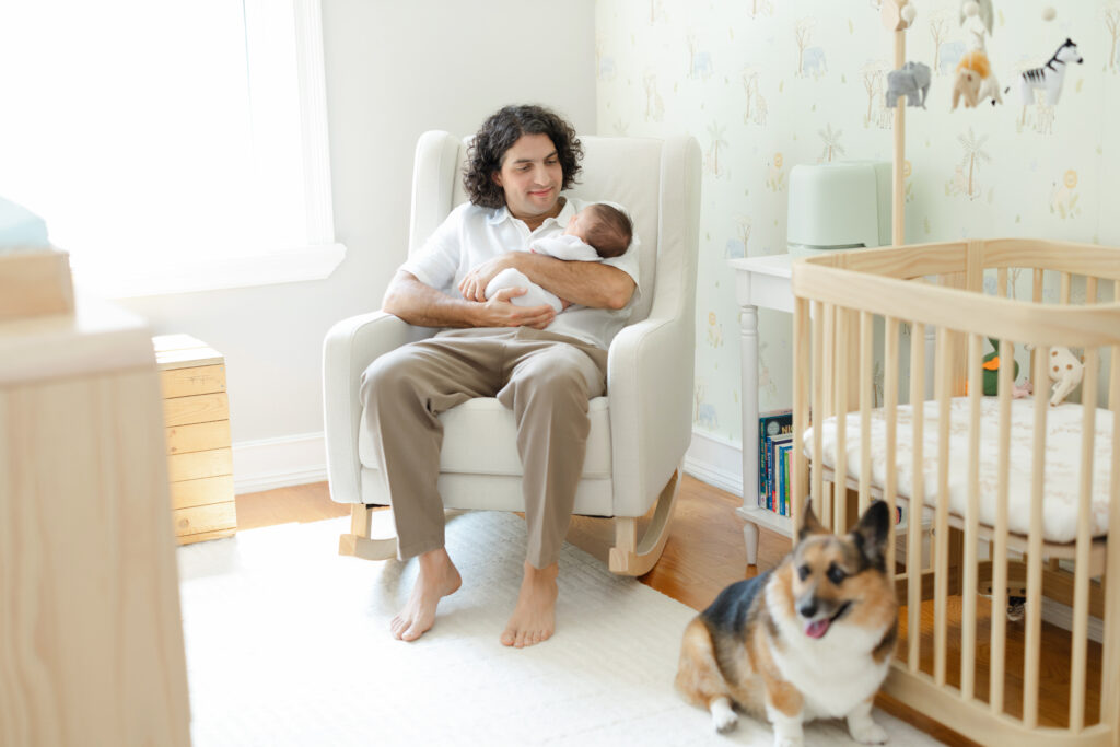 Father holding his newborn son in the nursery with the family corgi nearby