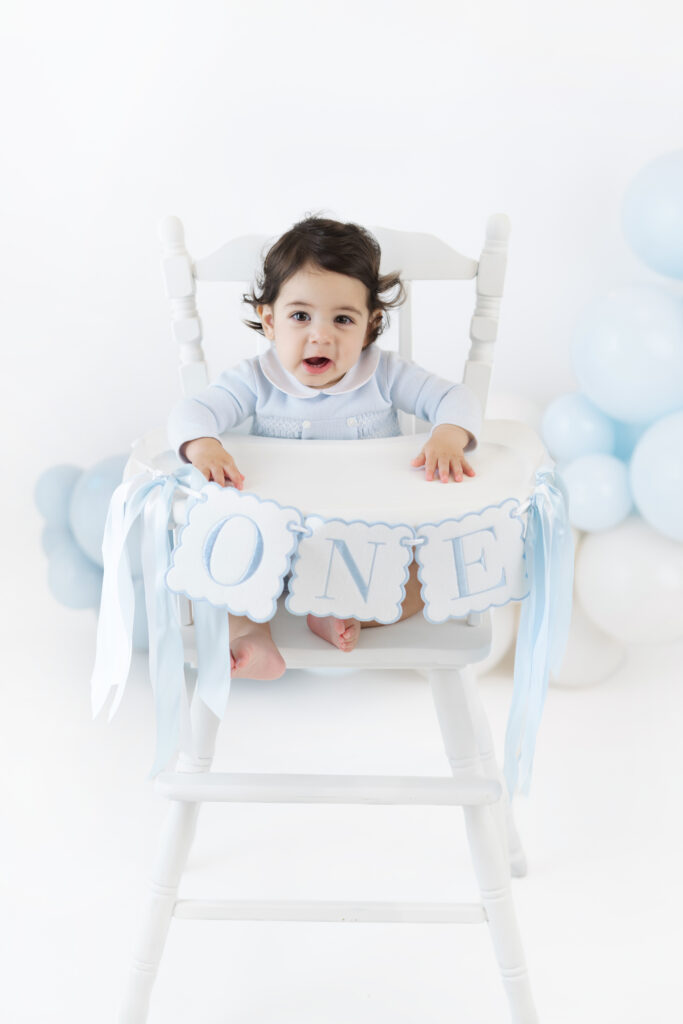 Soren in a white high chair with a first birthday banner and blue balloons